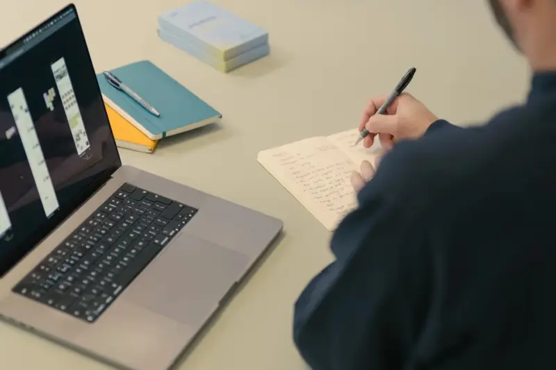 Person taking handwritten notes beside a laptop showing design screens