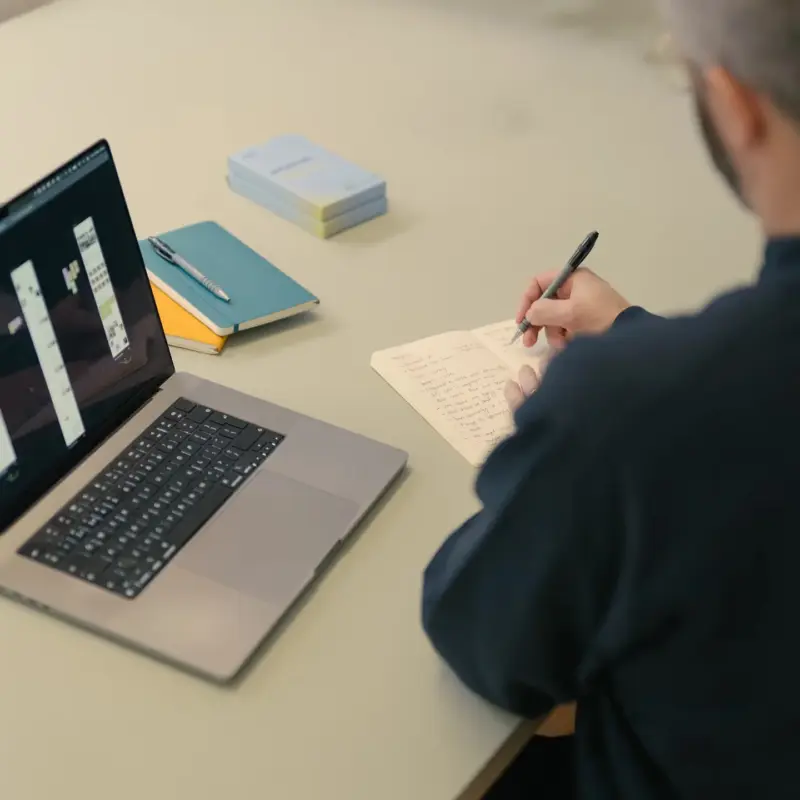 Person taking handwritten notes beside a laptop showing design screens