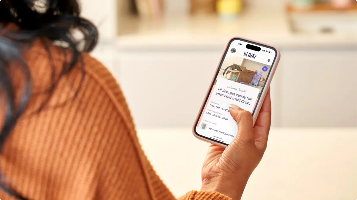 Woman holding a phone showing the Blink app welcome screen, 'Hi Joe, get ready for your next meal drop'