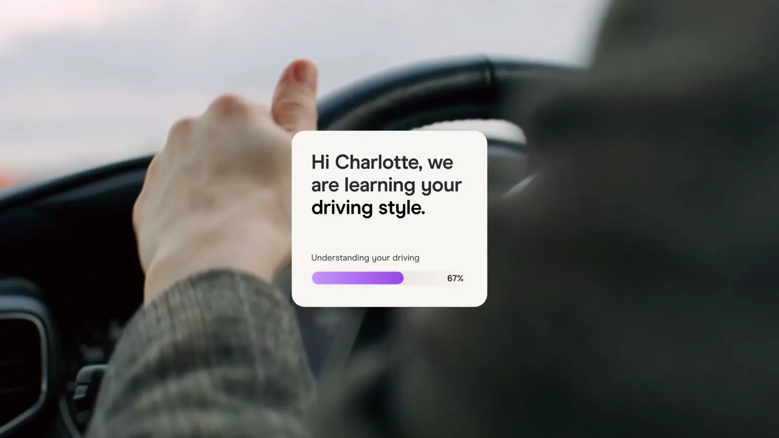 Driver's hand on a steering wheel with an app message, 'Hi Charlotte, we are learning your driving style'